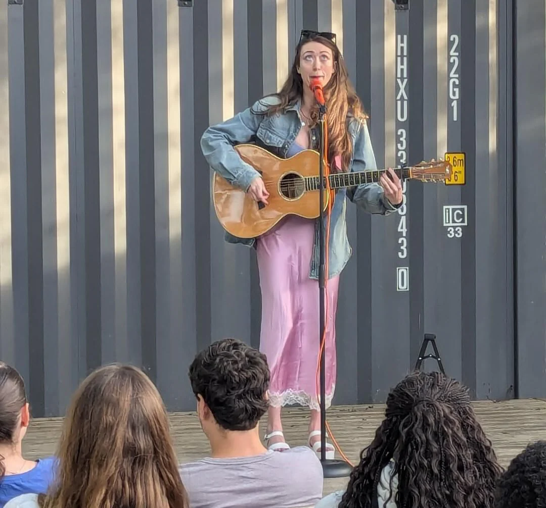 Sierra “Sisi” Moses performing an original comedy song on acoustic guitar for an outdoor crowd